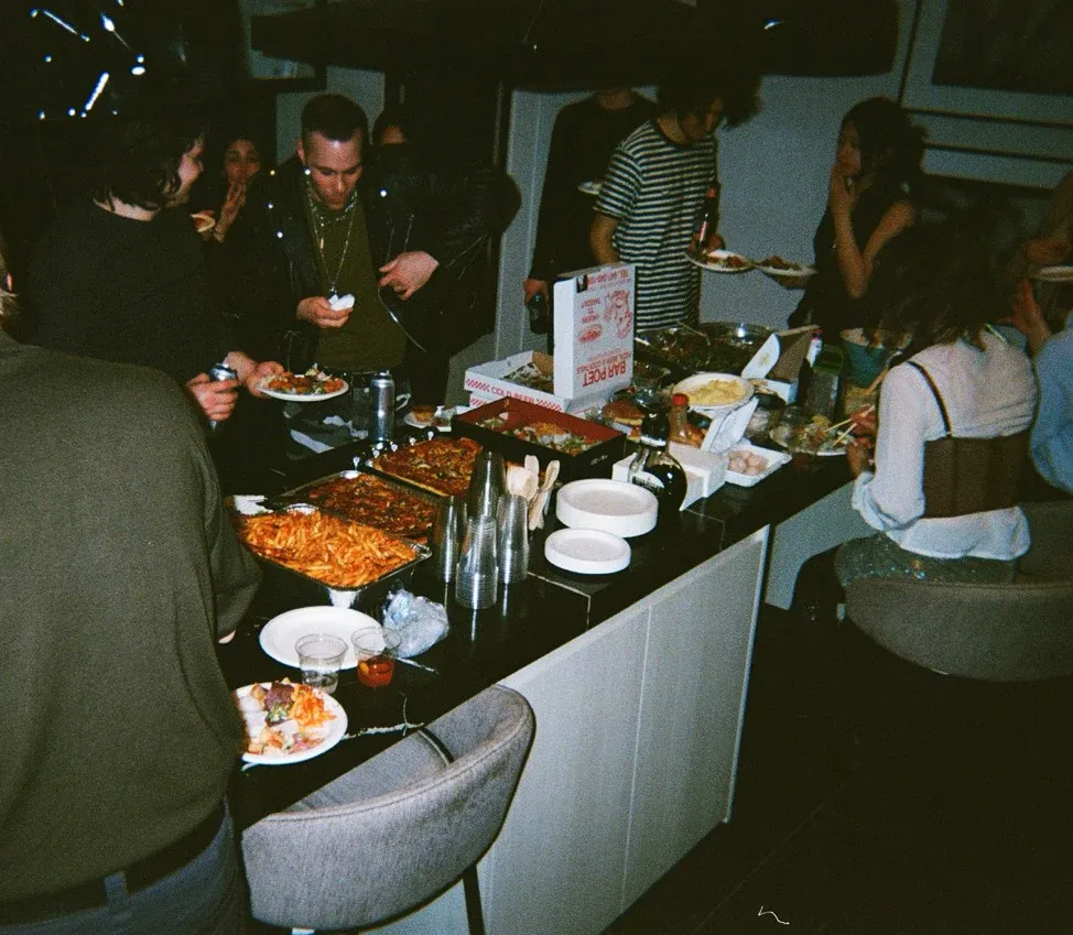 Wide shot of Fond dinner party with guests gathered around long kitchen counter talking and eating